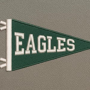 Puede incluir: Un banderín verde con la palabra "EAGLES" bordada en blanco. El banderín es triangular y tiene detalles blancos en el lado izquierdo. El fondo es un gris neutro texturizado.