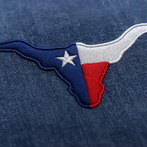 May include: Embroidered patch in the shape of Texas, featuring the state flag design. The patch is blue, white, and red, with a white star. The patch is sewn onto a blue denim fabric.