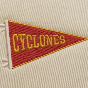 Peut inclure: Une bannière rouge avec le texte brodé en or "CYCLONES". La bannière a une bordure blanche et est placée sur un fond de tissu crème. La bannière est de forme triangulaire.