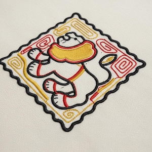 Op de afbeelding: Geborduurde patch met een gestileerde witte leeuw met rode en gele accenten, tegen een witte achtergrond. De leeuw bevindt zich in een ruitvormige rand met een zwarte geschulpte rand en decoratieve rode en gouden krullen.