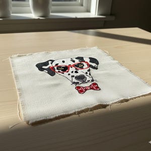 May include: Square fabric patch with an embroidered Dalmatian dog wearing red glasses and a red bow tie. The dog has black spots and is set against a white background with frayed edges. The patch is on a light-colored wooden surface.