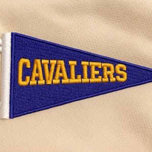 May include: A blue pennant with the word "CAVALIERS" embroidered in gold. The pennant has a white border and white fringe. The background is a light beige fabric, providing a neutral backdrop for the pennant.