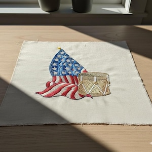 Könnte beinhalten: Gestickter Aufnäher mit einer amerikanischen Flagge und einer Trommel. Die Flagge hat blaue Sterne und rot-weiße Streifen. Die Trommel ist beige mit weißen Akzenten. Der Aufnäher befindet sich auf einem beigen Stoff.