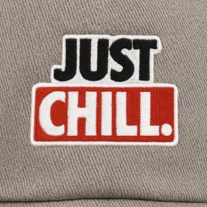 Op de afbeelding: Een grijze baseballpet met de geborduurde tekst "JUST CHILL". De tekst is in zwart en wit, met "CHILL" op een rode achtergrond. De pet is gemaakt van een textuurstof.