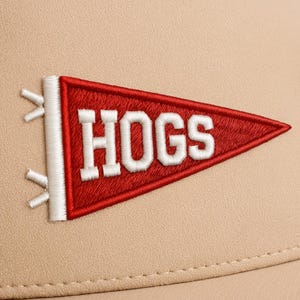May include: Close-up of a tan baseball cap with a red pennant-shaped patch. The patch features the word "HOGS" in white, embroidered lettering. The pennant has a white border and is attached to the cap with white stitching.