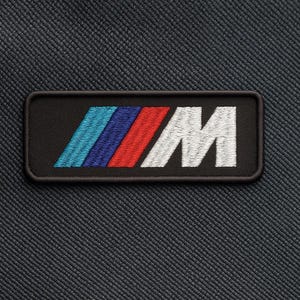 Pode incluir: Patch bordado com o logotipo da BMW M. O patch retangular tem um fundo preto com o logotipo M em azul, vermelho e branco. O patch está em um tecido texturizado cinza escuro.