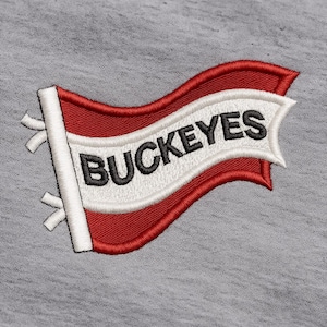Può includere: Toppa ricamata con un disegno a bandiera rossa e bianca con la scritta "BUCKEYES" in nero. La toppa è cucita su uno sfondo di tessuto grigio. La bandiera ha una forma ondulata.