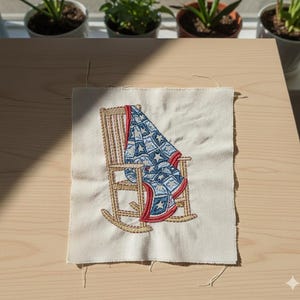 May include: Embroidered patch featuring a rocking chair with a blue and white quilt draped over it. The quilt has a star pattern and red trim. The chair is beige with gold stitching, set against a cream-colored background.