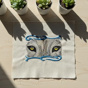May include: Embroidered patch with a wolf's eyes design. The eyes are yellow with black pupils, set in grey and white fur. A blue decorative border frames the eyes on a beige fabric square. Suitable for sewing or ironing onto clothing or accessories.