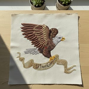 May include: Embroidered patch featuring a brown and white eagle with outstretched wings, clutching a banner that reads "Freedom" in gold thread. The eagle is set against a cream-colored fabric background, creating a patriotic theme.