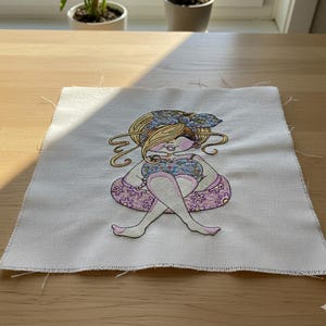 Pode incluir: Patch bordado com uma mulher sentada com cabelo loiro e um adorno floral na cabeça. A mulher usa um top floral e uma saia rosa com estampa floral. O patch está em um fundo de tecido branco.