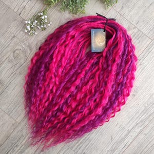 Pode incluir: Um conjunto de extensões de cabelo sintético com uma mistura vibrante de madeixas onduladas em rosa choque e roxo profundo. O cabelo está preso com um laço preto e inclui uma pequena etiqueta retangular com o texto The Moonlight Workshop.