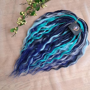 May include: Braided synthetic hair extensions in shades of blue, teal, and gray. The hair is styled with wavy ends and a tag attached. The extensions are arranged on a light brown surface.
