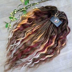 Puede incluir: Un conjunto de rastas onduladas multicolores. Las rastas presentan una mezcla de hebras marrones, rojas, rubias y blancas. Una etiqueta con el texto "THE MOONLIGHT DREADS" está adjunta. Las rastas están dispuestas sobre una superficie gris claro.