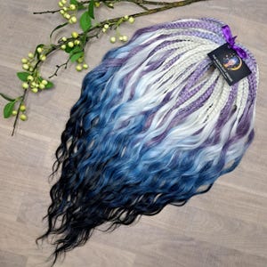 May include: Braided synthetic hair extensions in shades of blue, white, and purple, with a wavy texture. The extensions are arranged in a bundle, with a tag attached that reads "THE MOONLIGHT DREADS". A sprig of greenery with small, round, yellow-green berries is in the background.