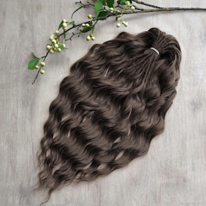 May include: Dark brown wavy hair extensions, styled with a small elastic band at the top. The extensions are arranged on a light wood-grain surface, with a sprig of greenery with small white berries.