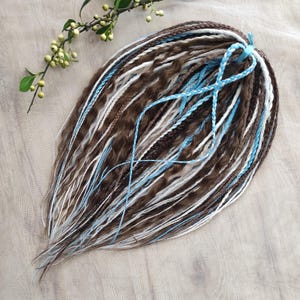 May include: A set of brown, white, and light blue dreadlocks. The dreadlocks are styled with a wavy texture and are tied together with a light blue ribbon. The dreadlocks are arranged on a light wood-grain surface.