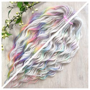 May include: A bundle of long, wavy, synthetic hair extensions in shades of grey, pastel rainbow, and white. The hair is tied with a pink ribbon. The extensions are styled to appear as dreadlocks. Green leaves and white berries are in the upper left corner.