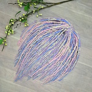 May include: A bundle of pastel-colored dreadlocks in shades of pink, blue, lavender, and cream. The dreadlocks are arranged in a circular shape, with a textured appearance. A sprig of greenery with small white berries is in the background.