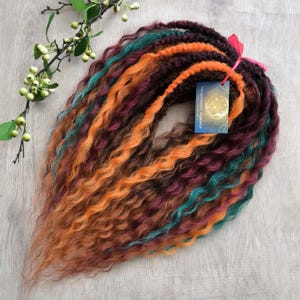 May include: A set of synthetic dreadlocks in a variety of colors, including teal, burgundy, orange, and brown. The dreadlocks are curly and are tied together with a pink ribbon. The label "The Moonlight Workshop" is attached to the dreadlocks.