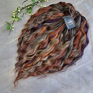 May include: A bundle of wavy, colorful hair extensions in shades of orange, purple, brown, and blonde. A tag with the text "The Moonlight Dreads" is attached with twine. A sprig of greenery with small white berries is in the upper left corner.