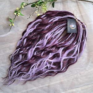 May include: A collection of burgundy and lilac synthetic dreadlocks with a wavy texture. The dreadlocks are tied with twine and a tag that reads "THE MOONLIGHT DREADS". A sprig of greenery with small white flowers is in the background.