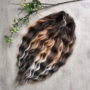 Puede incluir: Extensiones de cabello ondulado marrón oscuro con mechas rubias y melocotón. Las extensiones están peinadas con una ligera onda y atadas con hilo natural. Una ramita de vegetación con bayas blancas está en la esquina superior izquierda.