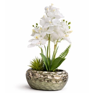 May include: An artificial orchid arrangement in a metallic silver-coloured pot. The arrangement features white orchid blooms, green leaves, and a small succulent plant. The pot has a textured, dimpled surface.