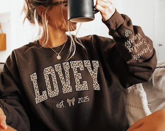 May include: Dark chocolate brown sweatshirt with the word "LOVEY" in a leopard print design. The sweatshirt also has the text "est. 2025" and names on the sleeve. A black mug is held up to the face.