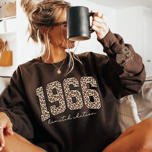 May include: A brown sweatshirt with the year "1966" in a leopard print design. The words "limited edition" are printed below the year. A black mug is held up to the face. The sleeve has a leopard print design with the words "limited edition".
