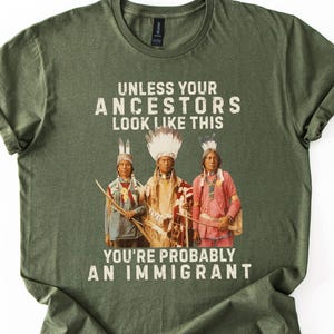 May include: Olive green t-shirt with the text "UNLESS YOUR ANCESTORS LOOK LIKE THIS YOU'RE PROBABLY AN IMMIGRANT." The graphic features three Native American figures in traditional attire. The shirt is made of a soft material.