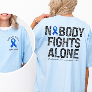 May include: Light blue t-shirt with the text "N&BODY FIGHTS ALONE" and a blue ribbon graphic on the back. The front of the shirt has a blue ribbon graphic and the words "SUPPORT SQUAD" and "TEAM JAMIE".