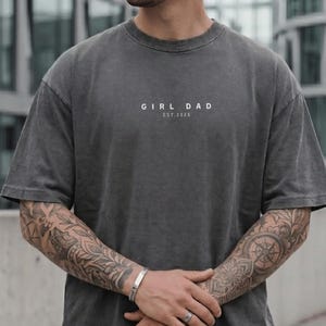 May include: Dark gray t-shirt with the text "GIRL DAD EST. 2026" in white. The shirt has short sleeves and a relaxed fit. The model is wearing a silver bracelet and ring.