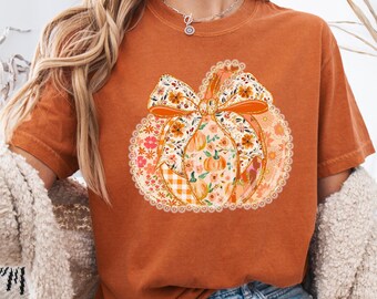 May include: Burnt orange t-shirt featuring a pumpkin design. The pumpkin is decorated with floral patterns, a bow, and a lace trim. The design incorporates orange, pink, and white colors. The shirt is short-sleeved and has a relaxed fit, perfect for fall.