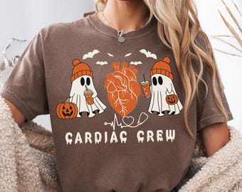 May include: Brown t-shirt with a Halloween-themed graphic. The design features two ghost characters wearing orange beanies and holding drinks, a pumpkin, and a stylized orange heart. The text "CARDIAC CREW" is printed below the graphic.