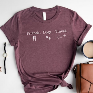May include: A maroon t-shirt with the words "Friends. Dogs. Travel." in white. The design includes silhouettes of people, paw prints, and an airplane trail. The shirt is knotted at the waist.