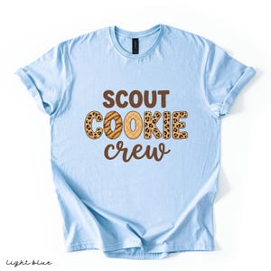 May include: Light blue t-shirt with the words "SCOUT COOKIE CREW" in brown and leopard print lettering. The shirt is a light blue color and has rolled-up sleeves. The words "light blue" are written in the bottom left corner.