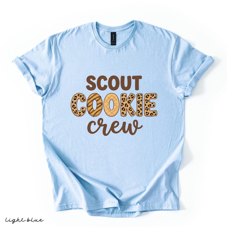 May include: Light blue t-shirt with the words "SCOUT COOKIE CREW" in brown and leopard print lettering. The shirt is a light blue color and has rolled-up sleeves. The words "light blue" are written in the bottom left corner.