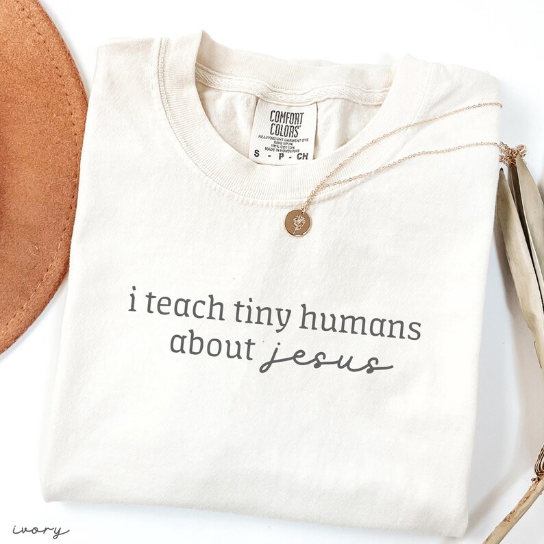 May include: Ivory-colored t-shirt with the text "i teach tiny humans about jesus" in dark gray. A gold necklace with a small pendant rests on the shirt. The shirt is folded and has a Comfort Colors tag.