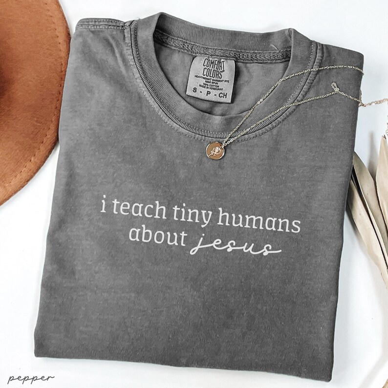 May include: A gray Comfort Colors t-shirt with the text "i teach tiny humans about jesus" in white script. A silver necklace with a small pendant rests on the shirt. A brown hat is partially visible in the upper left corner.