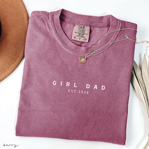 May include: A berry-colored Comfort Colors t-shirt with the text "GIRL DAD EST. 2026" in white. A gold necklace with a pendant rests on the shirt. The shirt is folded and has a tag at the neckline.