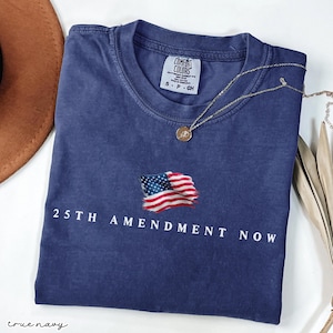 May include: A navy blue t-shirt with the text "25TH AMENDMENT NOW" and an embroidered American flag. The shirt is folded, with a gold necklace draped across it. The brand tag reads "Comfort Colors".