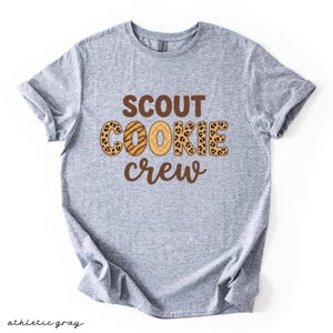 May include: Athletic gray t-shirt with the words "SCOUT COOKIE CREW" in brown and leopard print lettering. The shirt is made of a soft material and has rolled-up sleeves. The words "athletic gray" are written in cursive at the bottom.