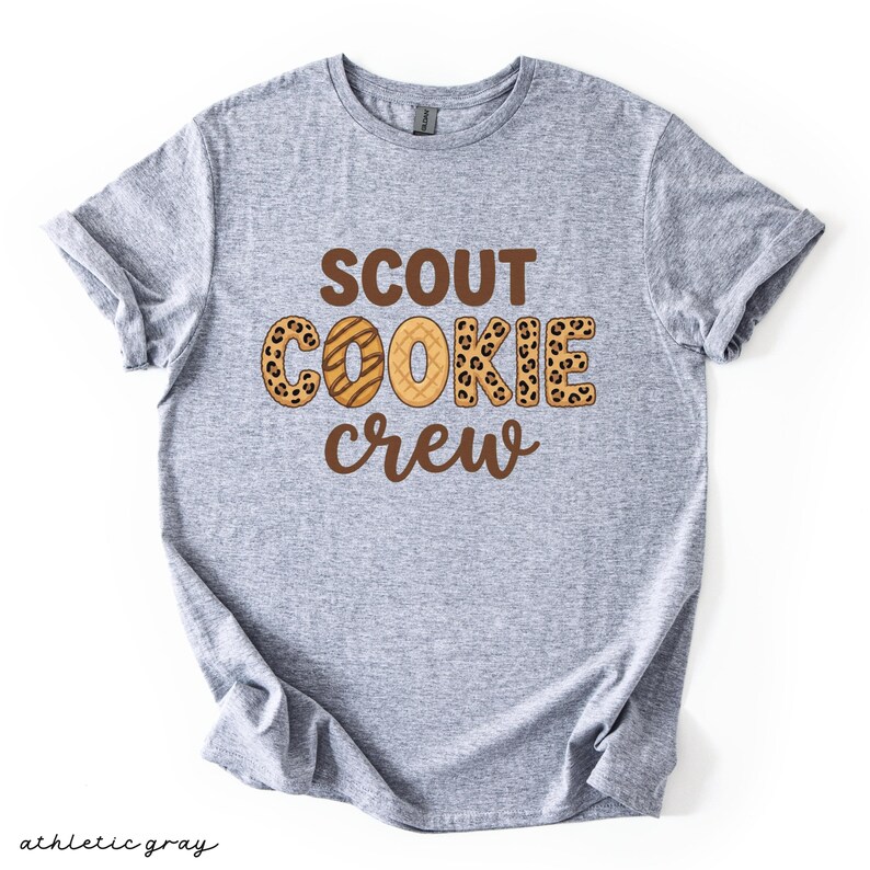 May include: Athletic gray t-shirt with the words "SCOUT COOKIE CREW" in brown and leopard print lettering. The shirt is made of a soft material and has rolled-up sleeves. The words "athletic gray" are written in cursive at the bottom.
