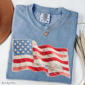 May include: A light blue Comfort Colors t-shirt with a sewn-on American flag design. The flag features white stars on a blue field and red and white stripes made of various patterned fabrics. A gold necklace with a pendant rests on the shirt.