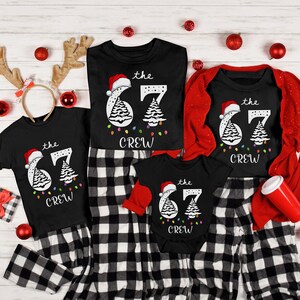May include: Black t-shirts and a onesie with matching black and white plaid pajama pants. Each shirt features a Christmas tree design with the text "the 67 crew". A red Santa hat is on the number 6. Christmas ornaments and reindeer antlers are scattered around.
