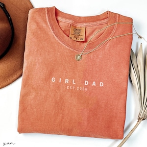 May include: A coral-colored t-shirt with the words "GIRL DAD EST. 2026" printed in white. A gold necklace with a pendant rests on the shirt. A brown hat and dried plant are in the background.