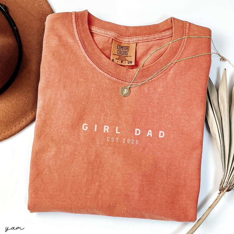 May include: A coral-colored t-shirt with the words "GIRL DAD EST. 2026" printed in white. A gold necklace with a pendant rests on the shirt. A brown hat and dried plant are in the background.