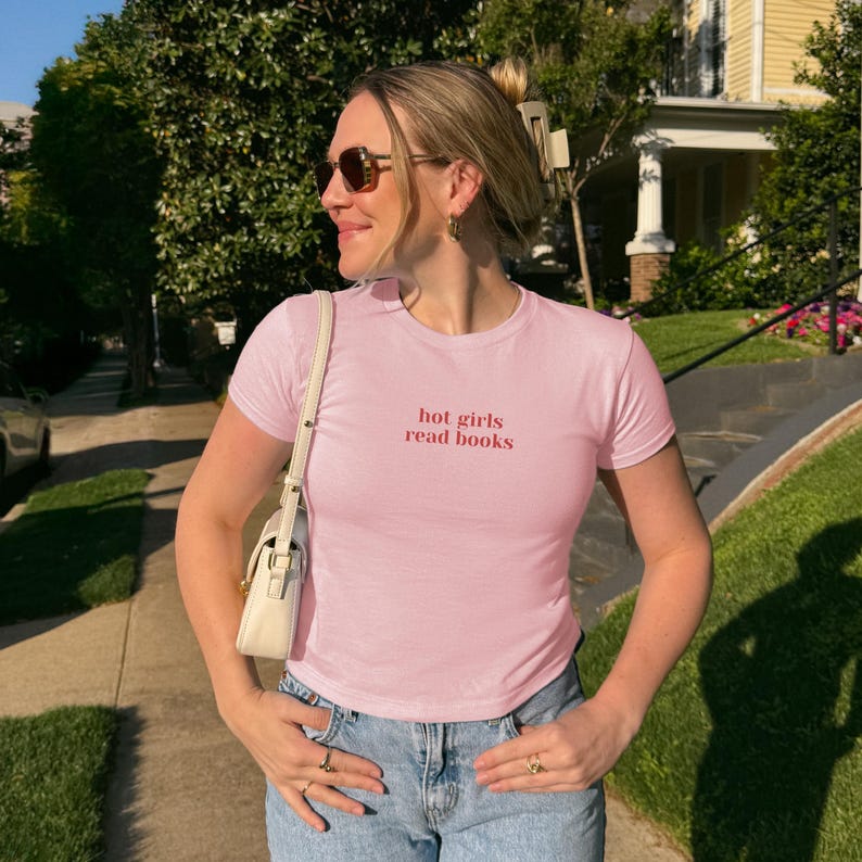 May include: A light pink t-shirt with the text "hot girls read books" in red. The shirt is paired with blue jeans and a white purse. The person is wearing sunglasses and rings.
