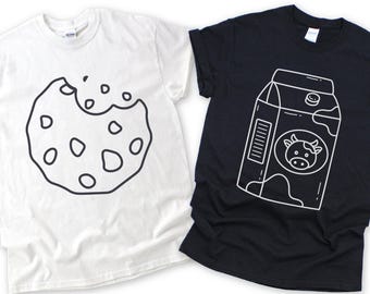 May include: Two t-shirts: one white, one black. The white shirt features a black outline of a bitten cookie. The black shirt displays a white outline of a milk carton with a cow graphic. Both shirts are short-sleeved.